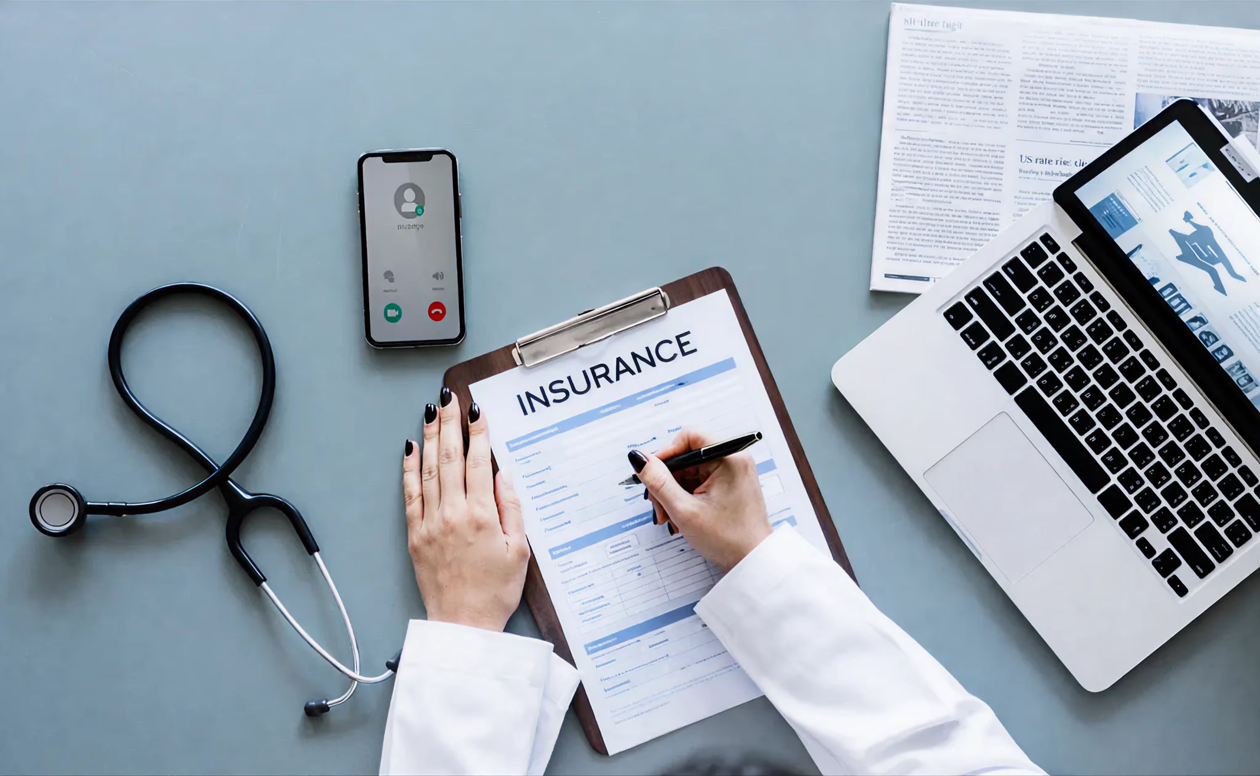 Hands filling out an insurance form on a clipboard beside a laptop and stethoscope